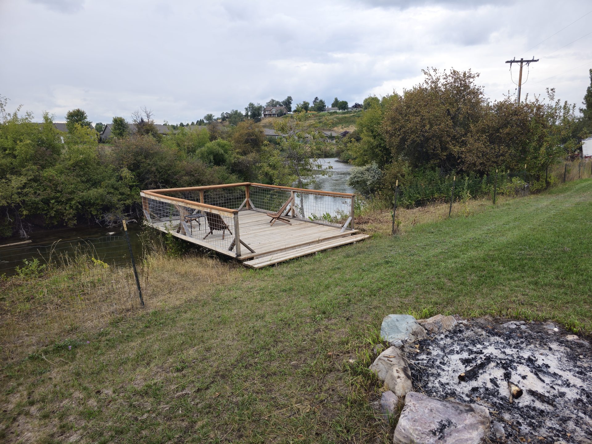 A riverside deck with chairs and a fire pit beside the Whitefish River in late summer